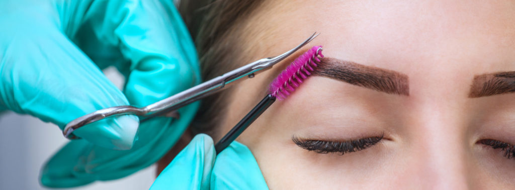 A technician using a small brush and long thin scissors to meticulously sculpt eyebrows.