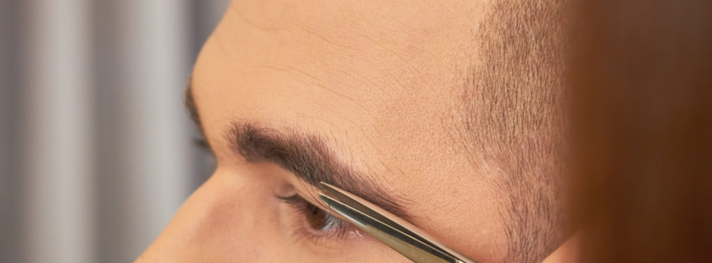 A man is getting his eyebrows trimmed by a pair of scissors, adding definition and shape.