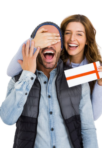 Beautiful smiling women covering her boyfriend's eyes as she surprises him with a gift certificate for laser hair removal.