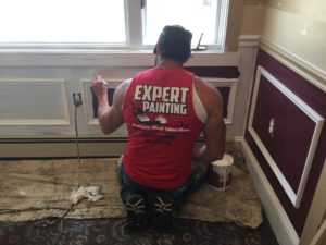 A man in a red shirt reading "Expert Painting" applies a coa of paint on the baseboards and lower walls.