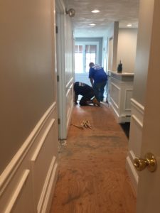 Two men at the end of a hallway working on the floor. One man kneels working, as another leans over assisting. 