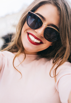 A woman with long flowing brown hair with softer brown hair, with sculpted eyebrows, sunglasses and vibrant smile.