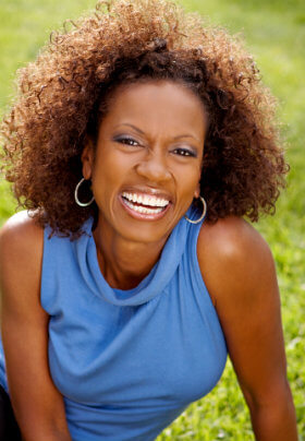 Woman with blue sleeveless blouse lounging on a grassy area with a large bright smile and bouncy hair.