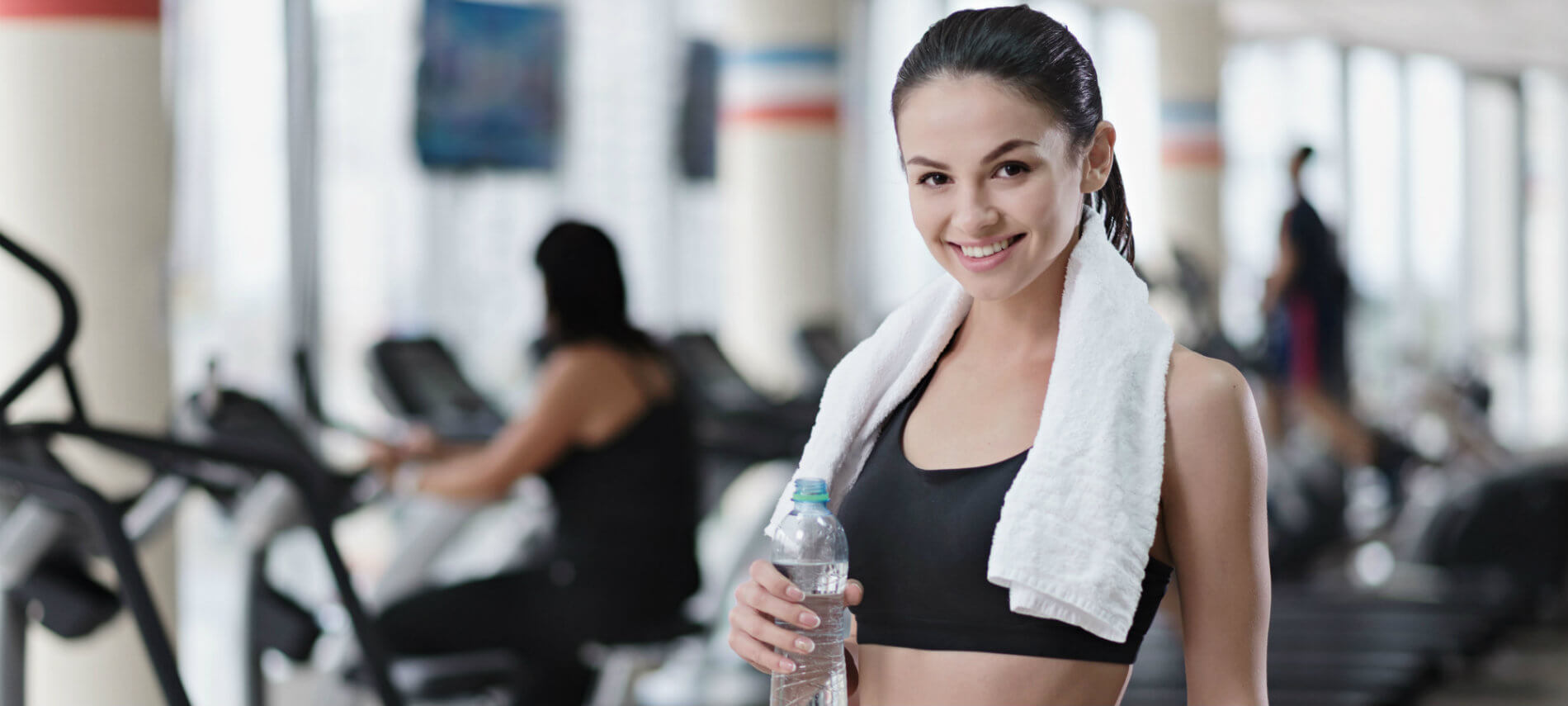 A woman in the gym with a towl around her shoulders pausing to take a drink.
