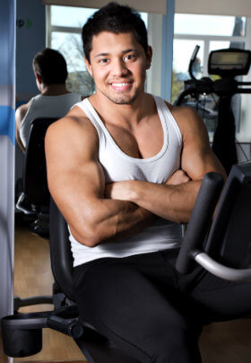 A man in shite muscle shirt sits on a piece of equipment with mirror in background posing/flexing his arms slightly in front of his smooth pecs.